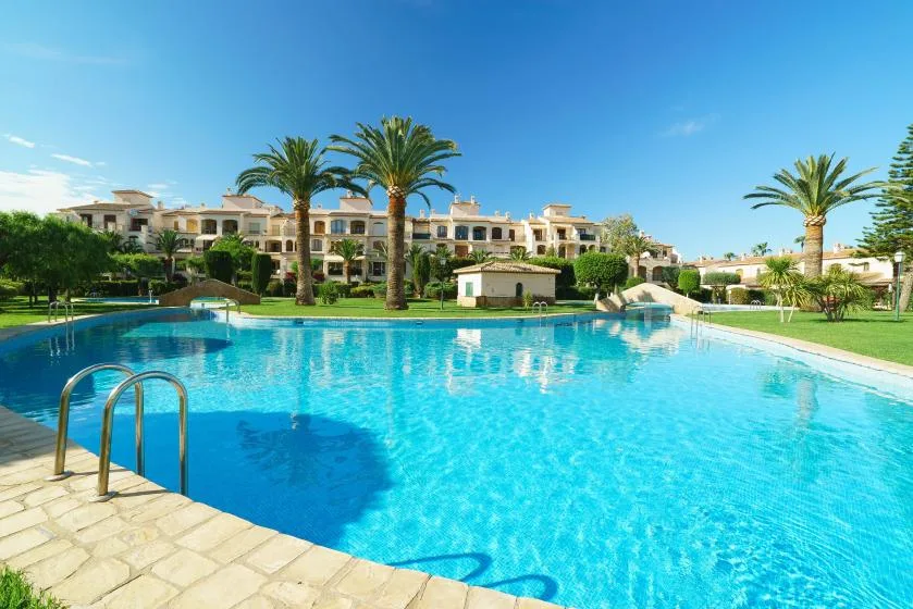 Una gran piscina al aire libre está rodeada de palmeras y edificios bajos.