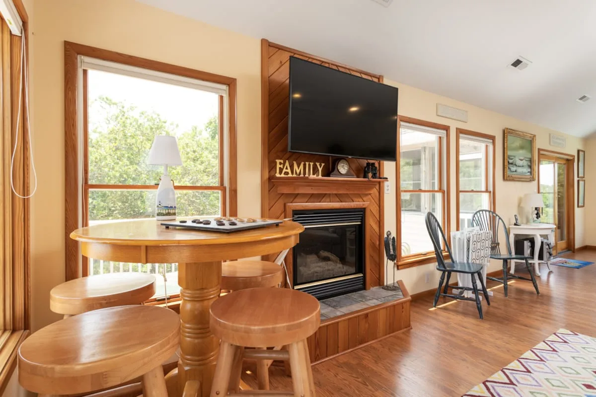 A bright living room with a wooden dining table and a television above the fireplace.