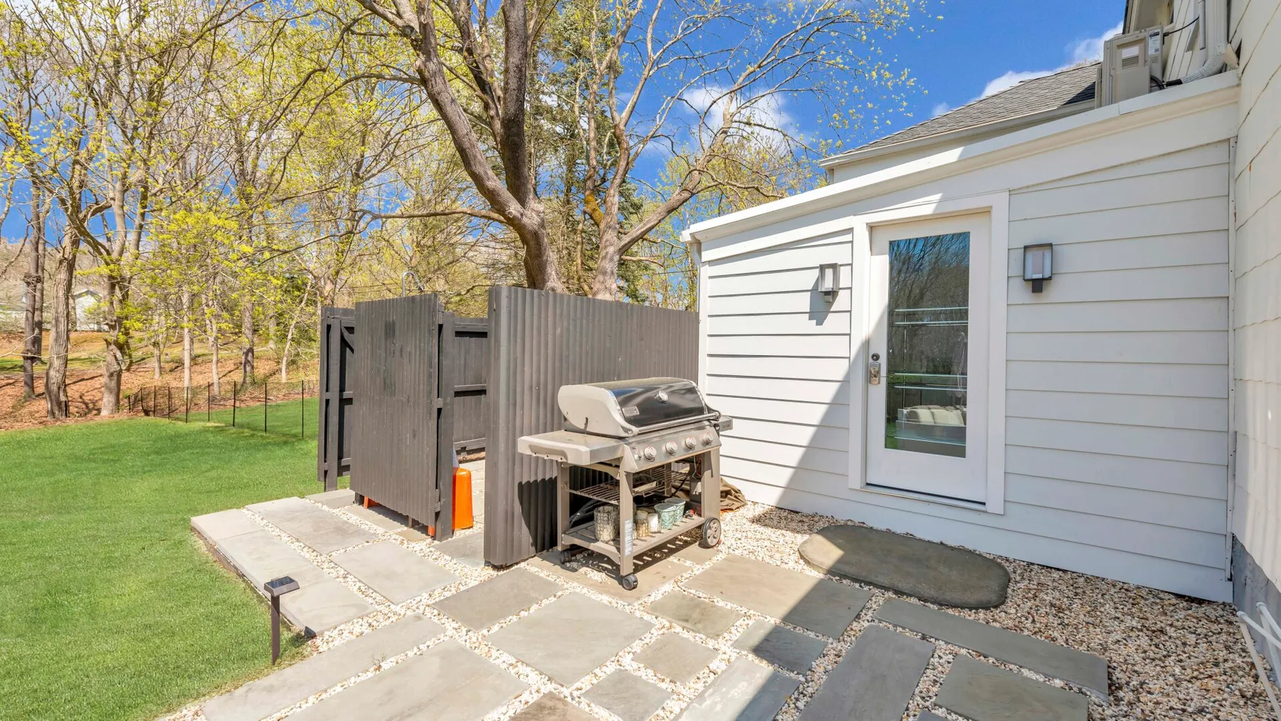 A paved outdoor patio beside a white-sided building, with a grill and fenced yard.