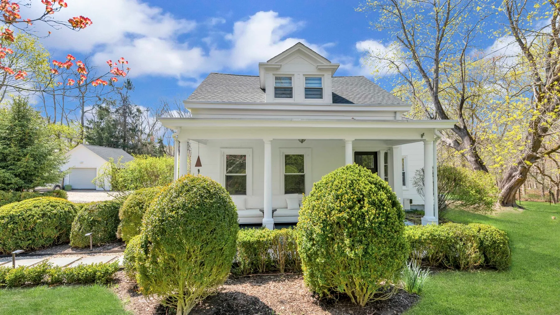 A white house with a covered front porch sits among shrubs and trees.