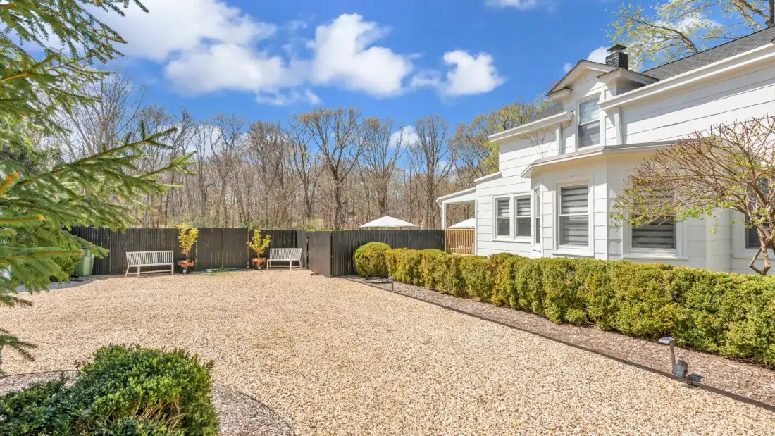 A gravel backyard beside a white house, bordered by hedges and a fence.