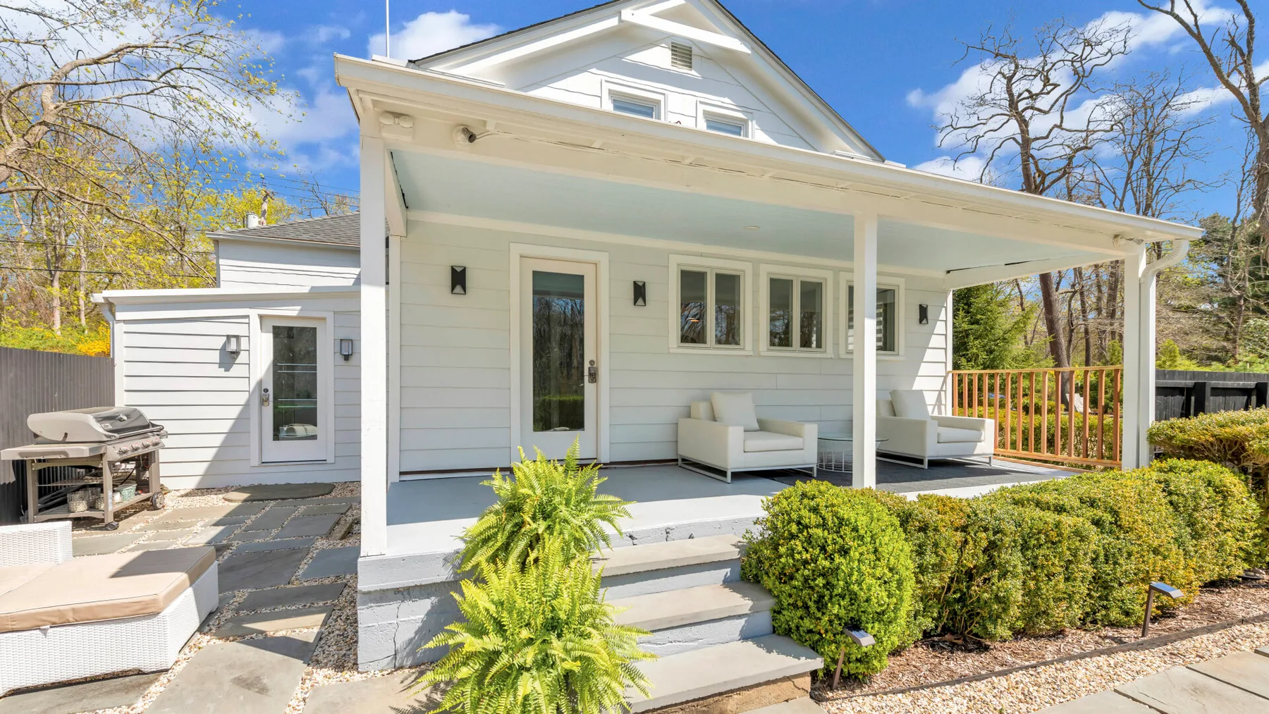 A white house with a covered porch, outdoor seating, and shrubs along the steps.