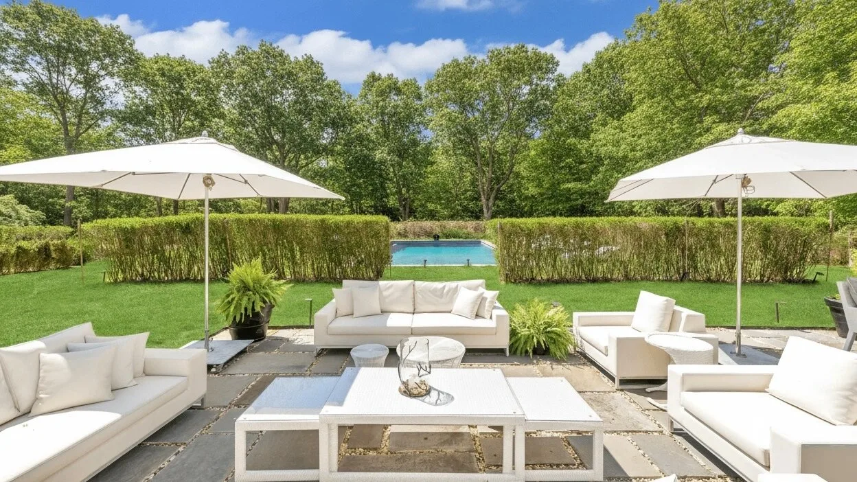 Outdoor patio seating surrounds a low table, with umbrellas overlooking a lawn and pool.