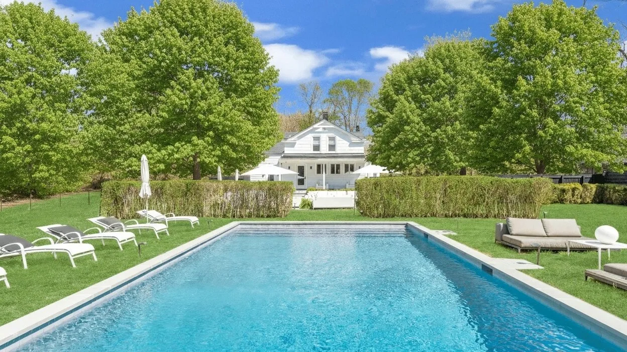 An outdoor swimming pool sits in a landscaped yard with lounge seating and a house beyond.