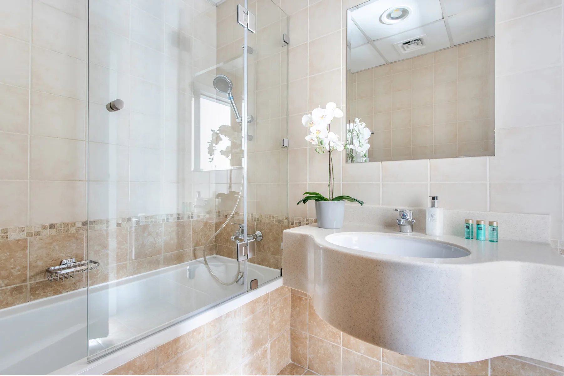 Un baño luminoso con bañera, mampara de ducha de vidrio y orquídea blanca.