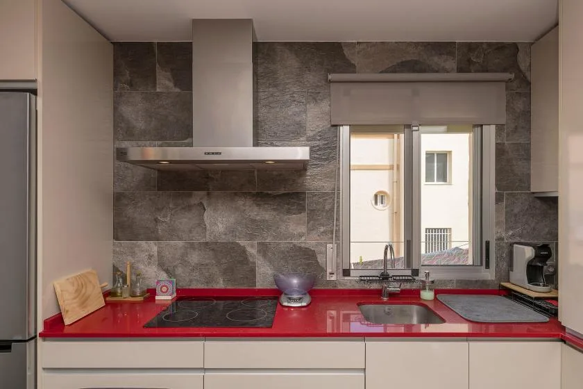 A modern kitchen with a red countertop, dark backsplash, and window above the sink.