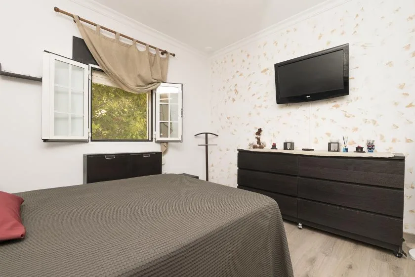 A bedroom with a bed, open windows, and a wall-mounted television.