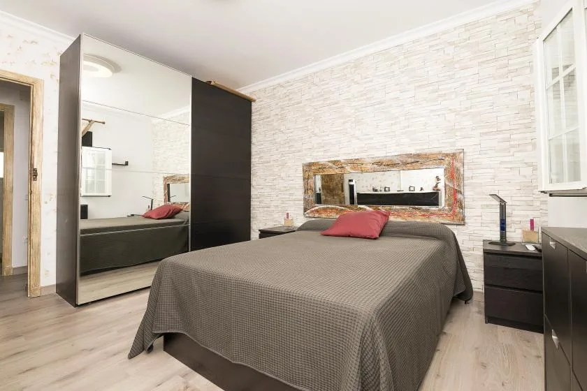 A bedroom with a bed and mirrored wardrobe against a light brick-patterned wall.