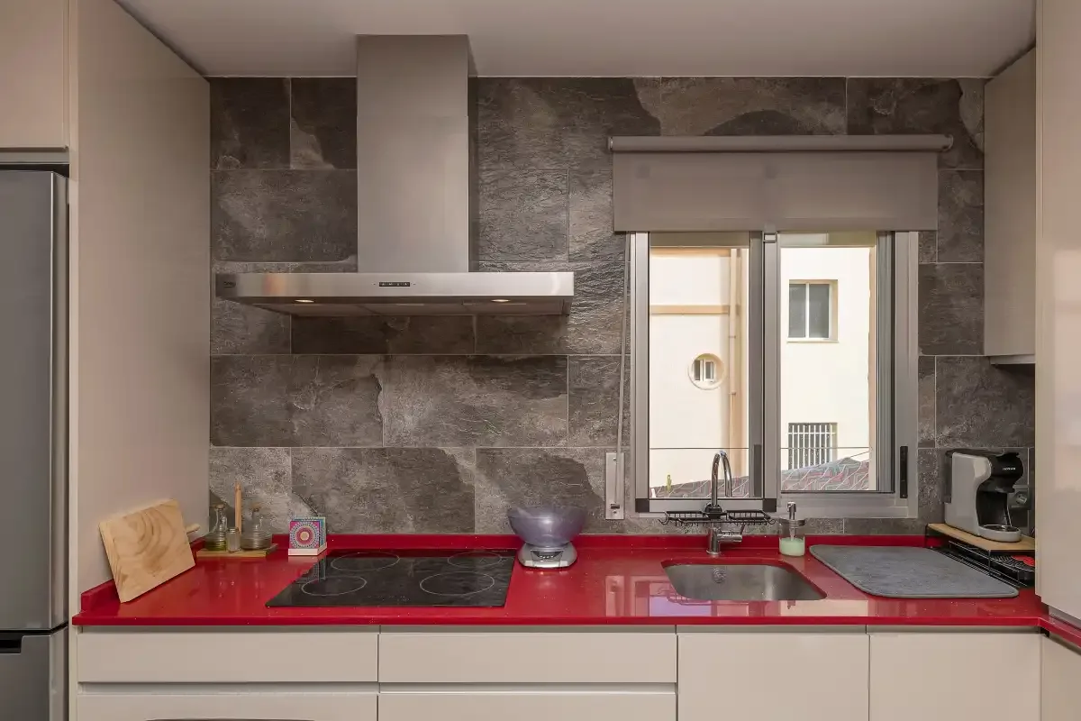 A modern kitchen with a red countertop, cooktop, and sink beneath a window.