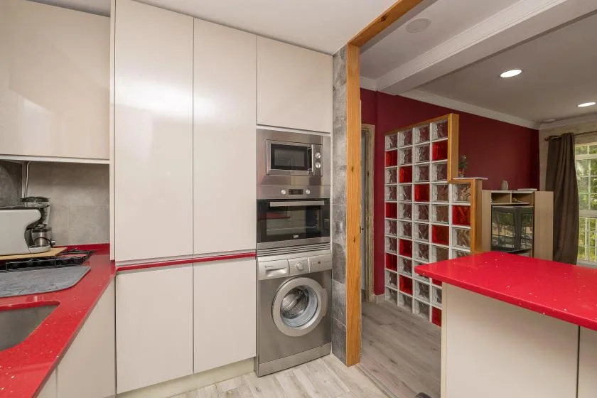 A modern kitchen with red countertops and built-in appliances opens onto a living area.