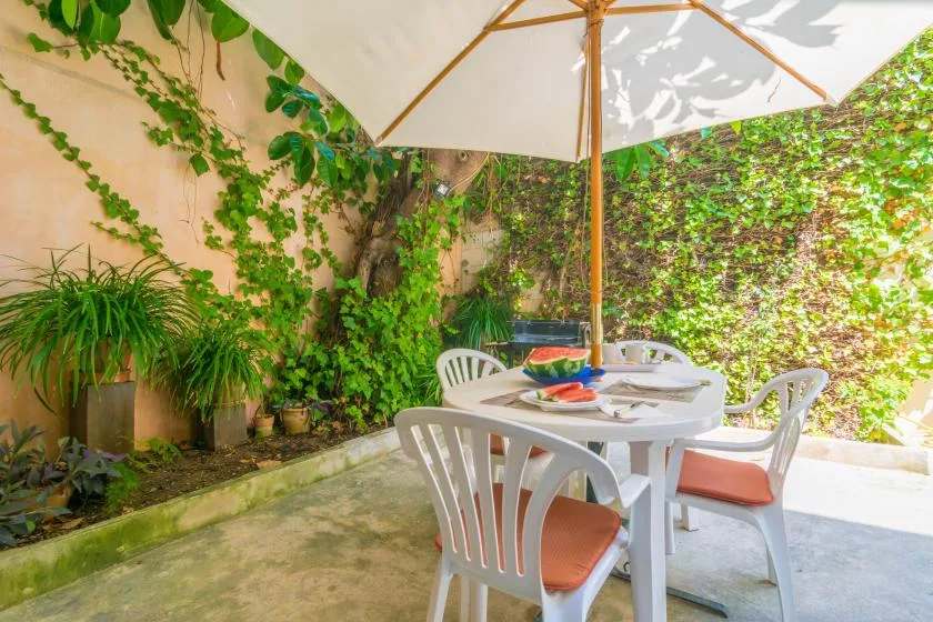 A patio dining area with white seating beneath a large umbrella, surrounded by greenery.
