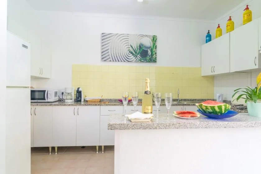 A bright kitchen with white cabinets, a counter, and a dining island set with glasses and fruit.