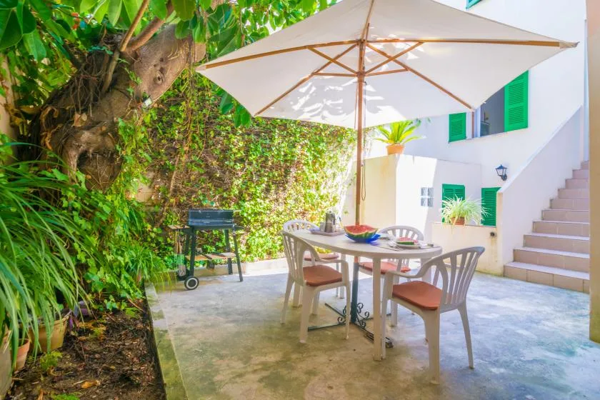 An outdoor patio dining area sits beneath a large umbrella beside leafy plants.