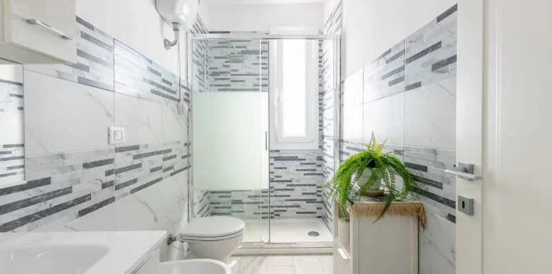 Un baño luminoso con azulejos estampados, ducha, lavabo, inodoro y una planta en maceta.
