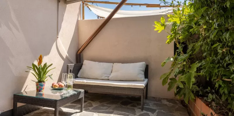 Una pequeña zona exterior para sentarse con cojines, una mesa baja y plantas en macetas.