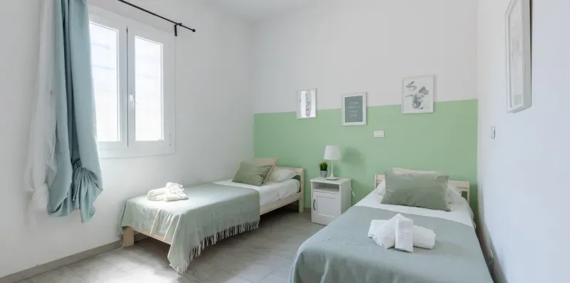 Un dormitorio luminoso con dos camas, paredes verde claro y una ventana grande.