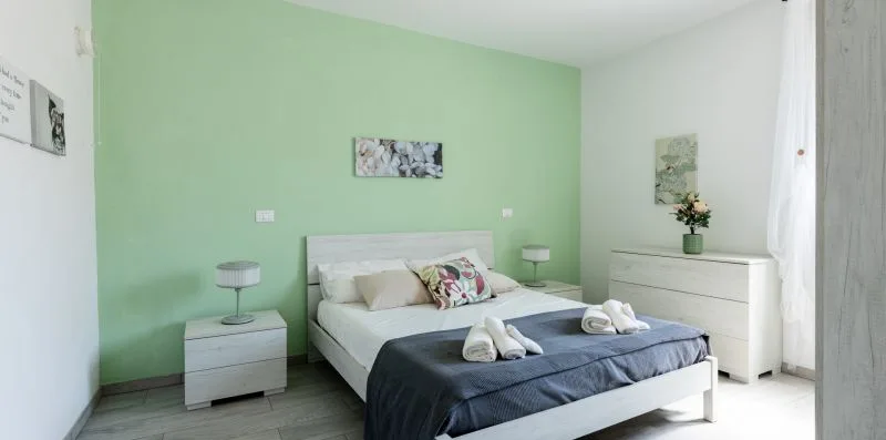 Un dormitorio con una cama vestida con ropa de cama blanca y azul oscuro frente a una pared verde claro.