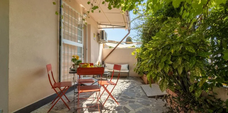 Un patio exterior sombreado con una mesa, sillas y plantas frondosas.