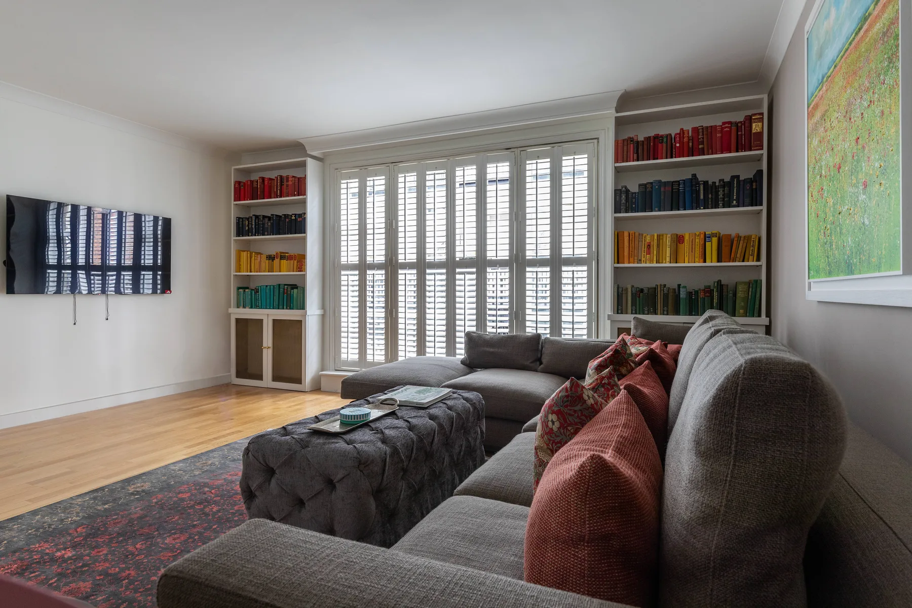 A living room with gray seating, a tufted ottoman, and bookshelves beside bright windows.