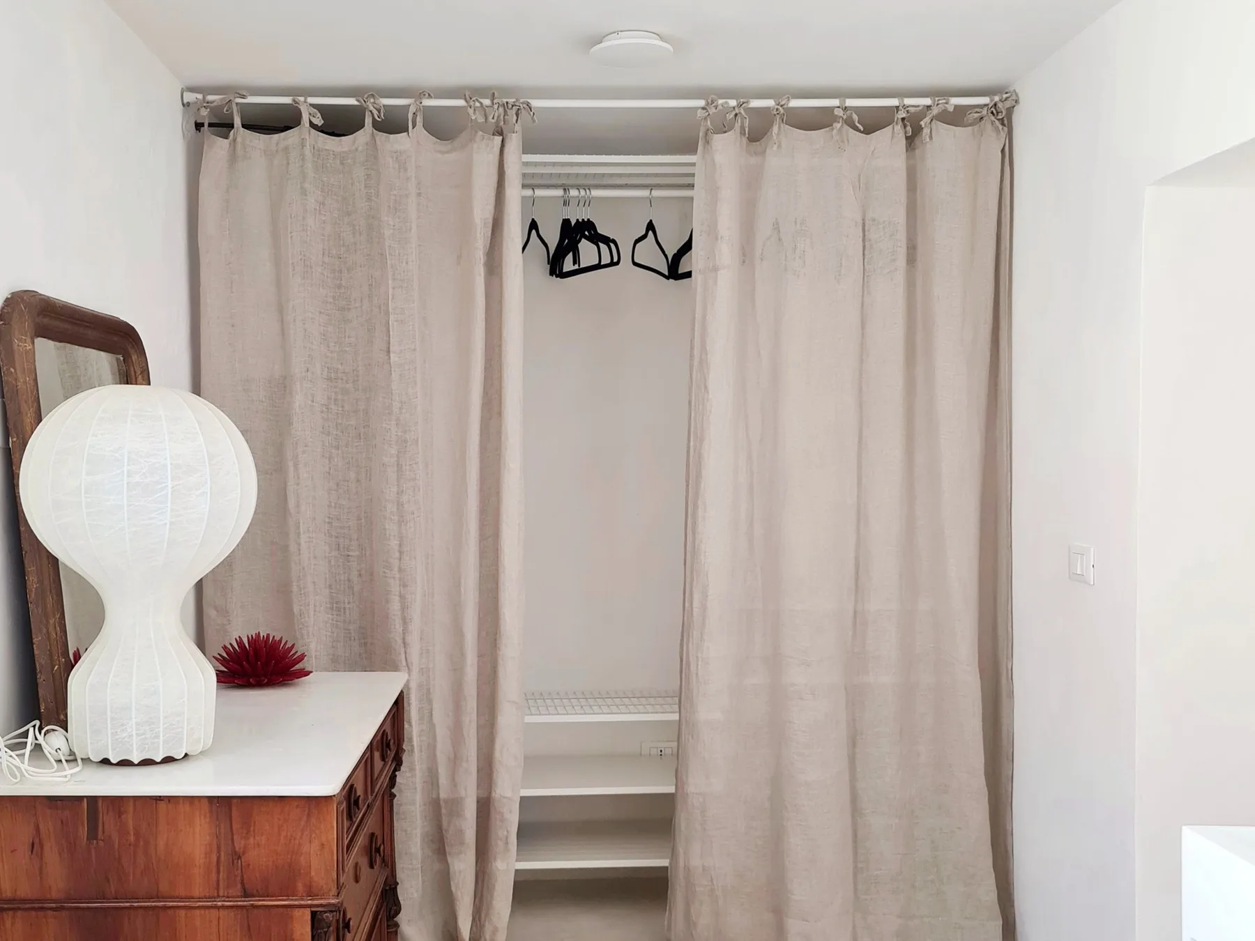 Un armario con cortinas, ropa colgada y una cómoda con una lámpara encima.