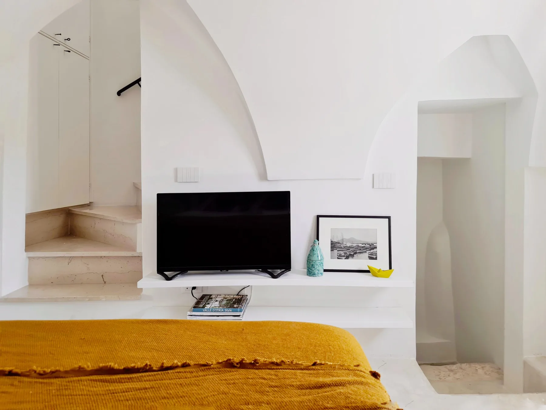 Un dormitorio con un televisor sobre un estante blanco bajo, detrás de una cama.