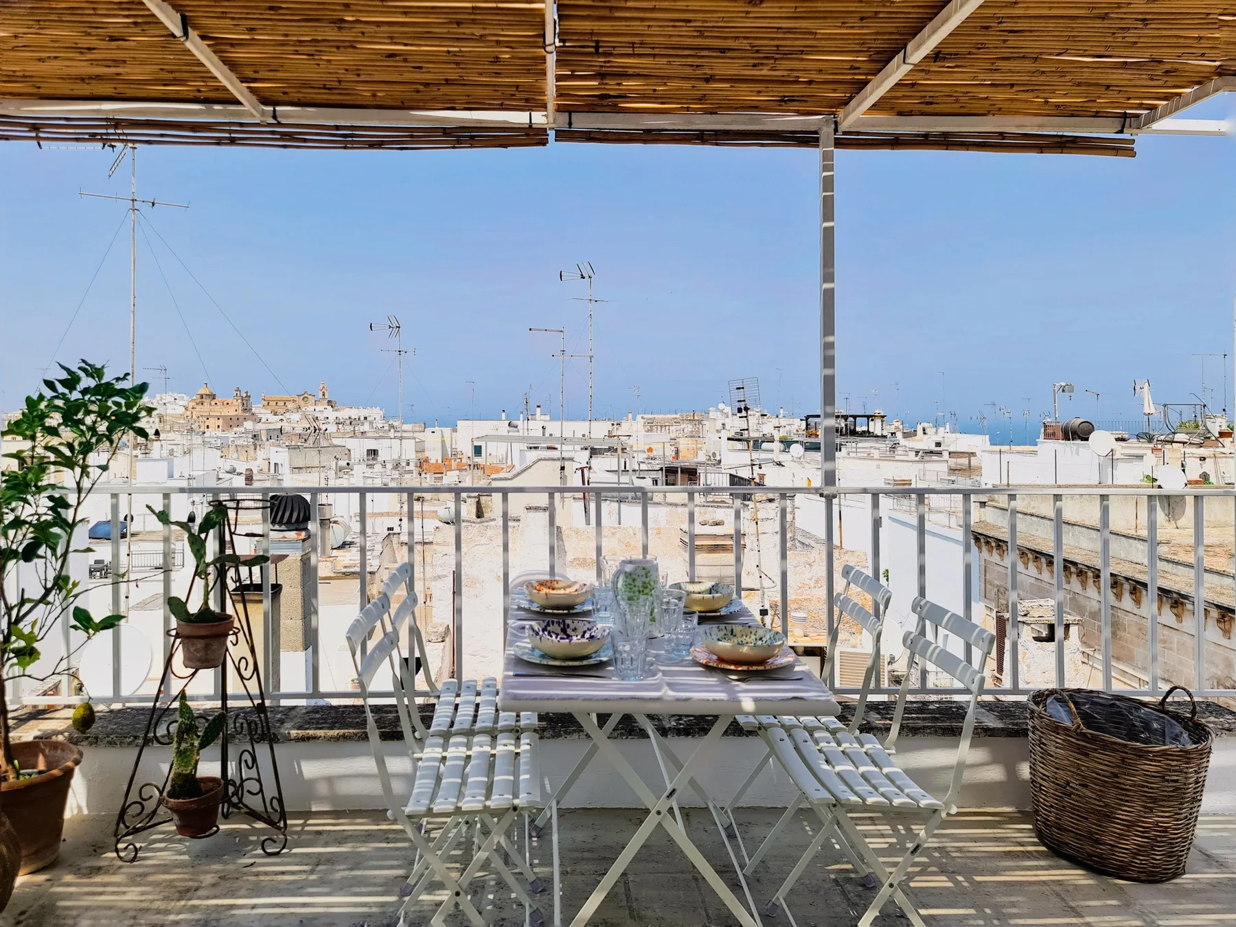 Una terraza sombreada tiene una mesa de comedor con vistas a edificios cercanos y al mar.