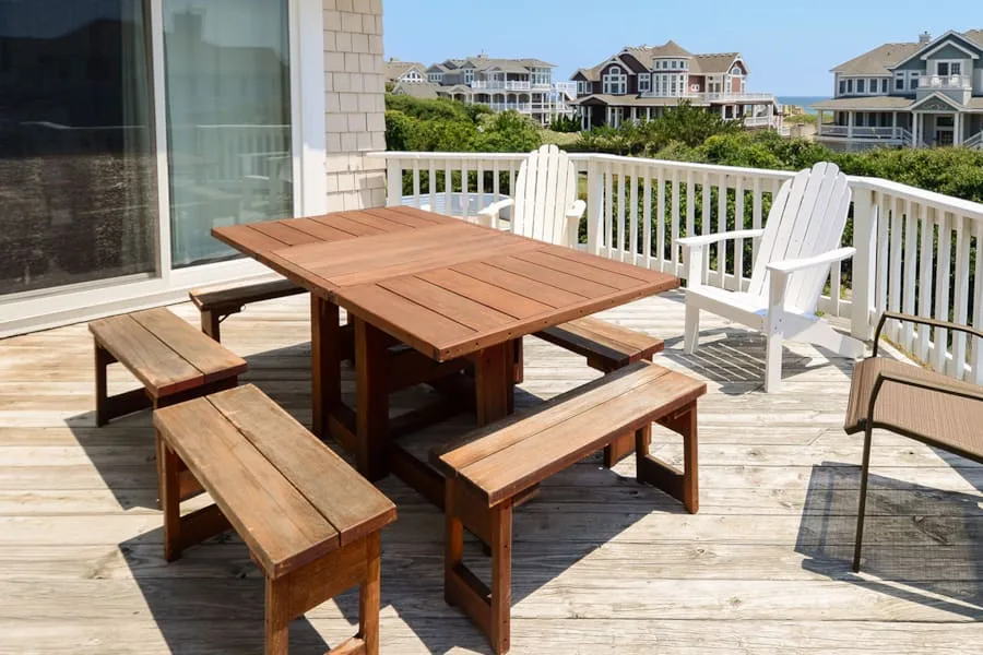 A wooden picnic table and benches sit on a sunny deck overlooking nearby houses.