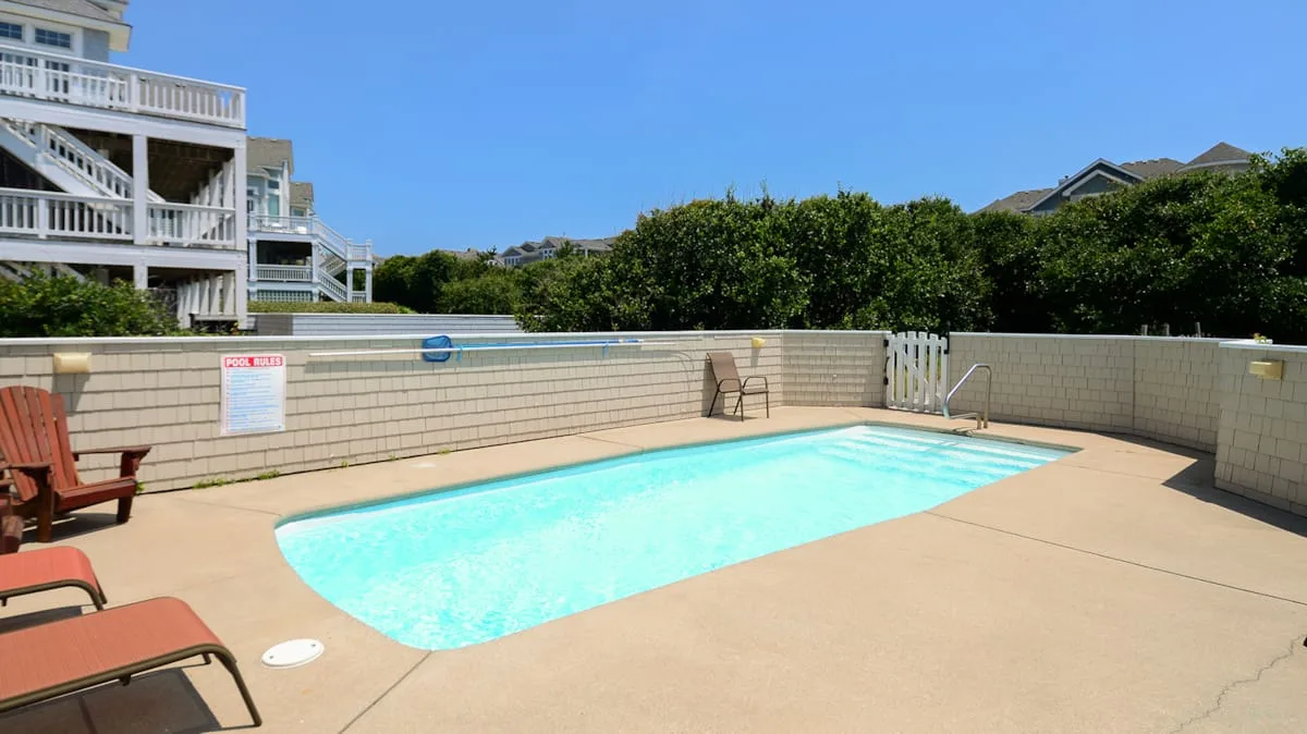 An outdoor swimming pool sits in a sunny paved patio beside a low wall.
