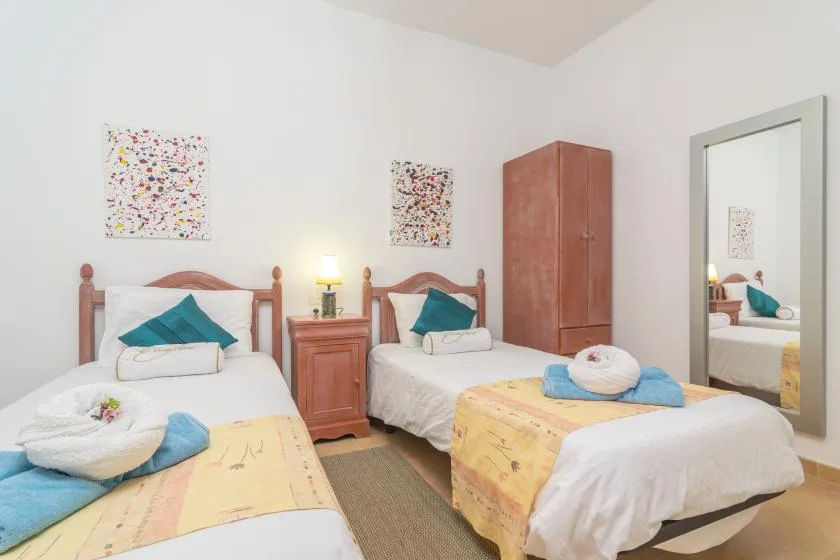 A twin bedroom with two single beds, colorful wall art, and a wooden wardrobe.