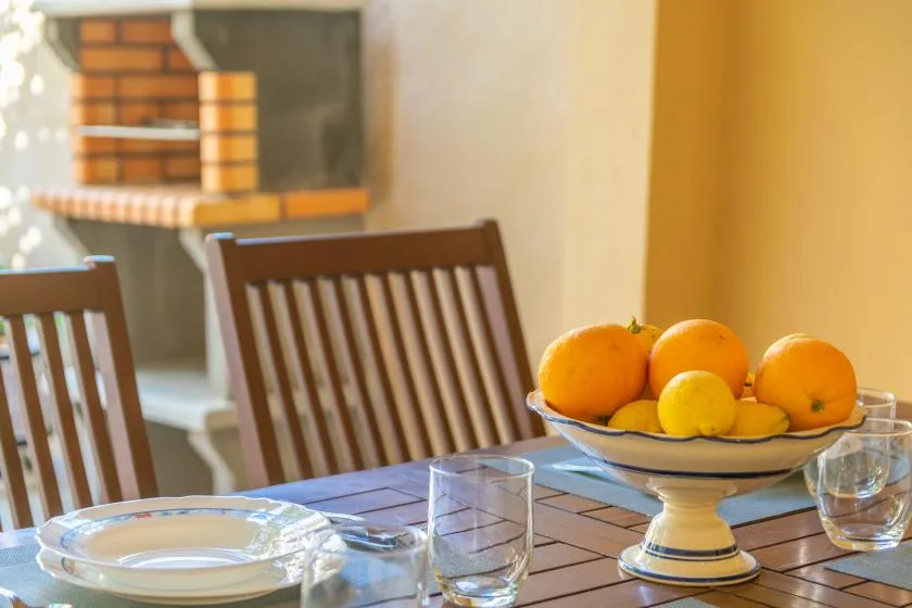 A dining table with place settings and a bowl of citrus fruit sits beside wooden seating.