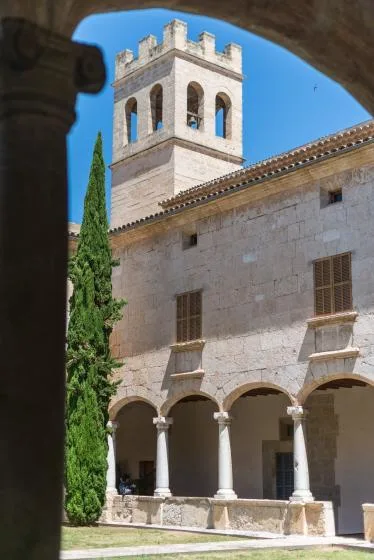 A stone courtyard with an arched walkway, a tall bell tower, and a cypress tree.