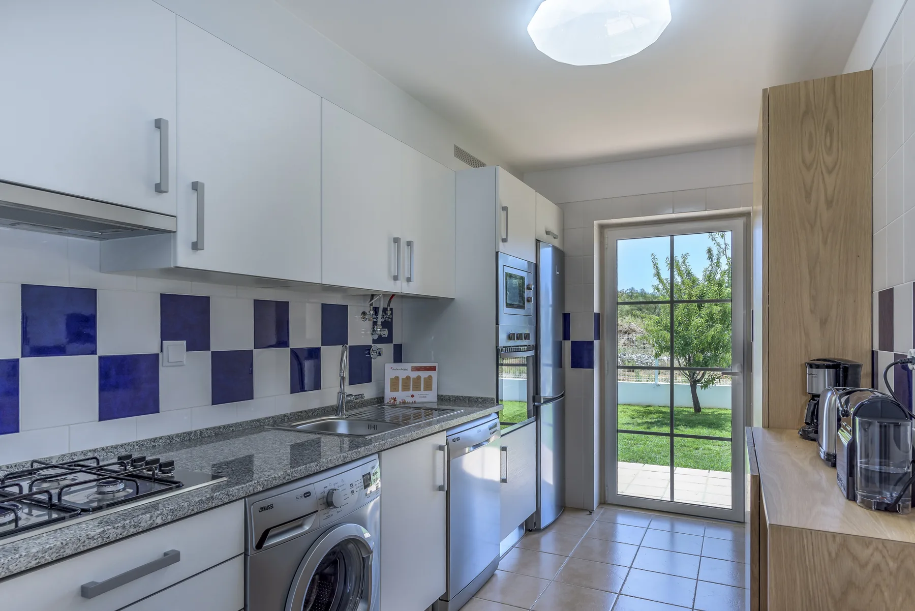 Una cocina luminosa con armarios blancos, encimeras de azulejos y una puerta acristalada al jardín.