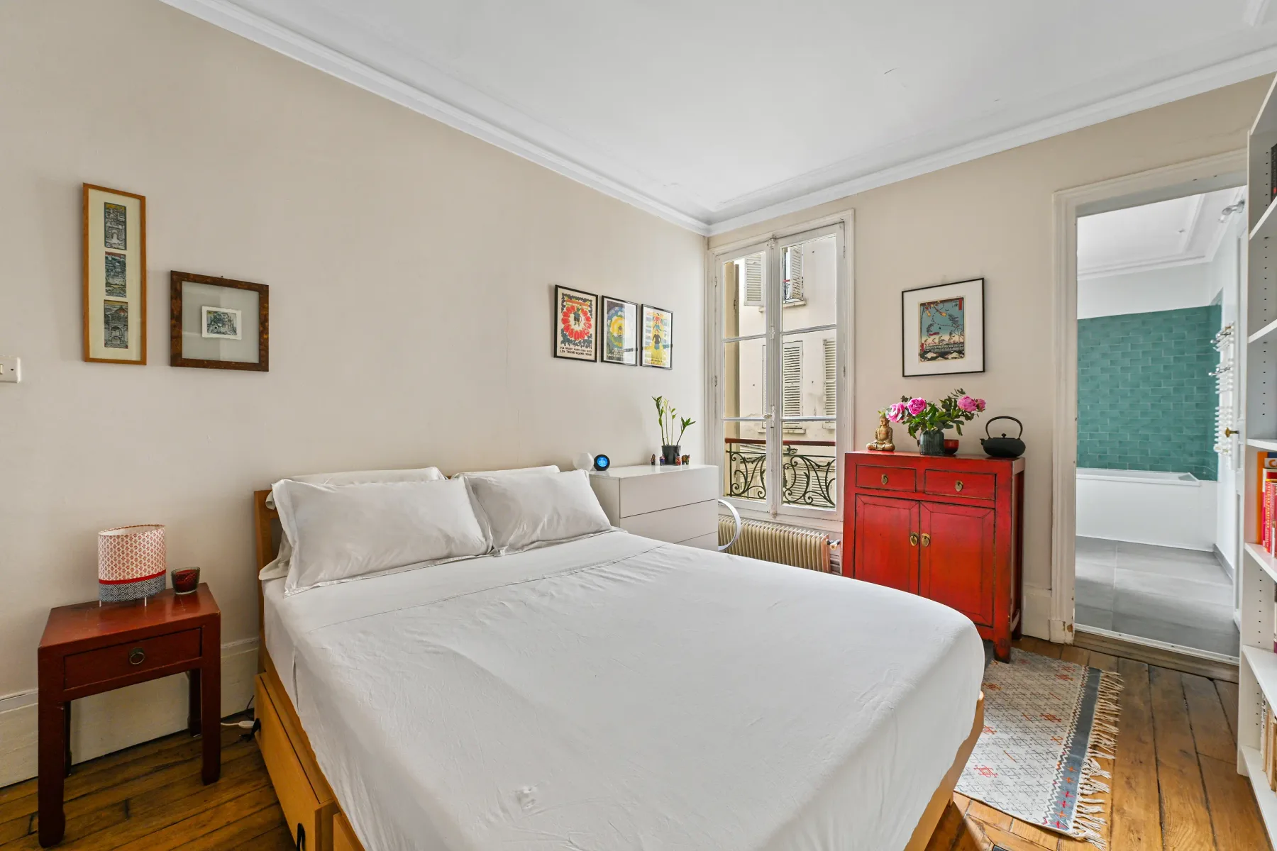 Un dormitorio con una cama tendida, cuadros enmarcados y una cómoda roja.