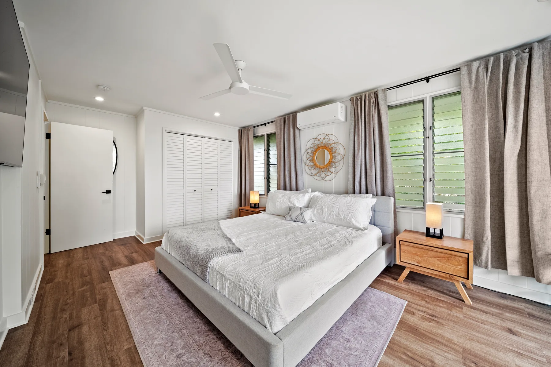 Un dormitorio luminoso tiene una cama grande junto a ventanas altas con cortinas.