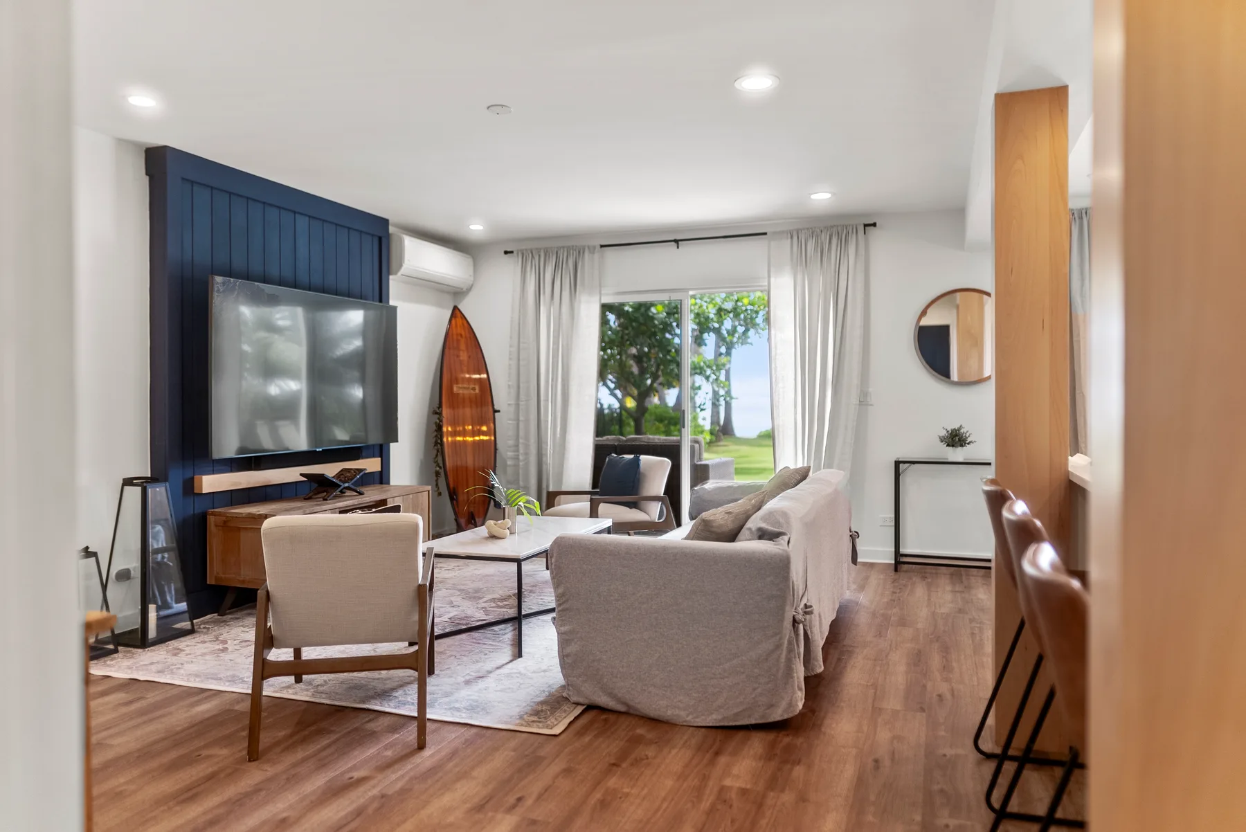 Sala de estar con asientos, un televisor y grandes ventanales con vista a la vegetación.