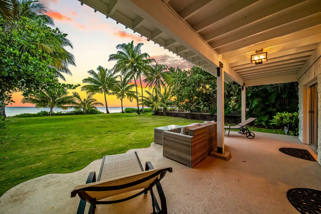 Un patio cubierto da a un césped verde, palmeras y el agua al atardecer.