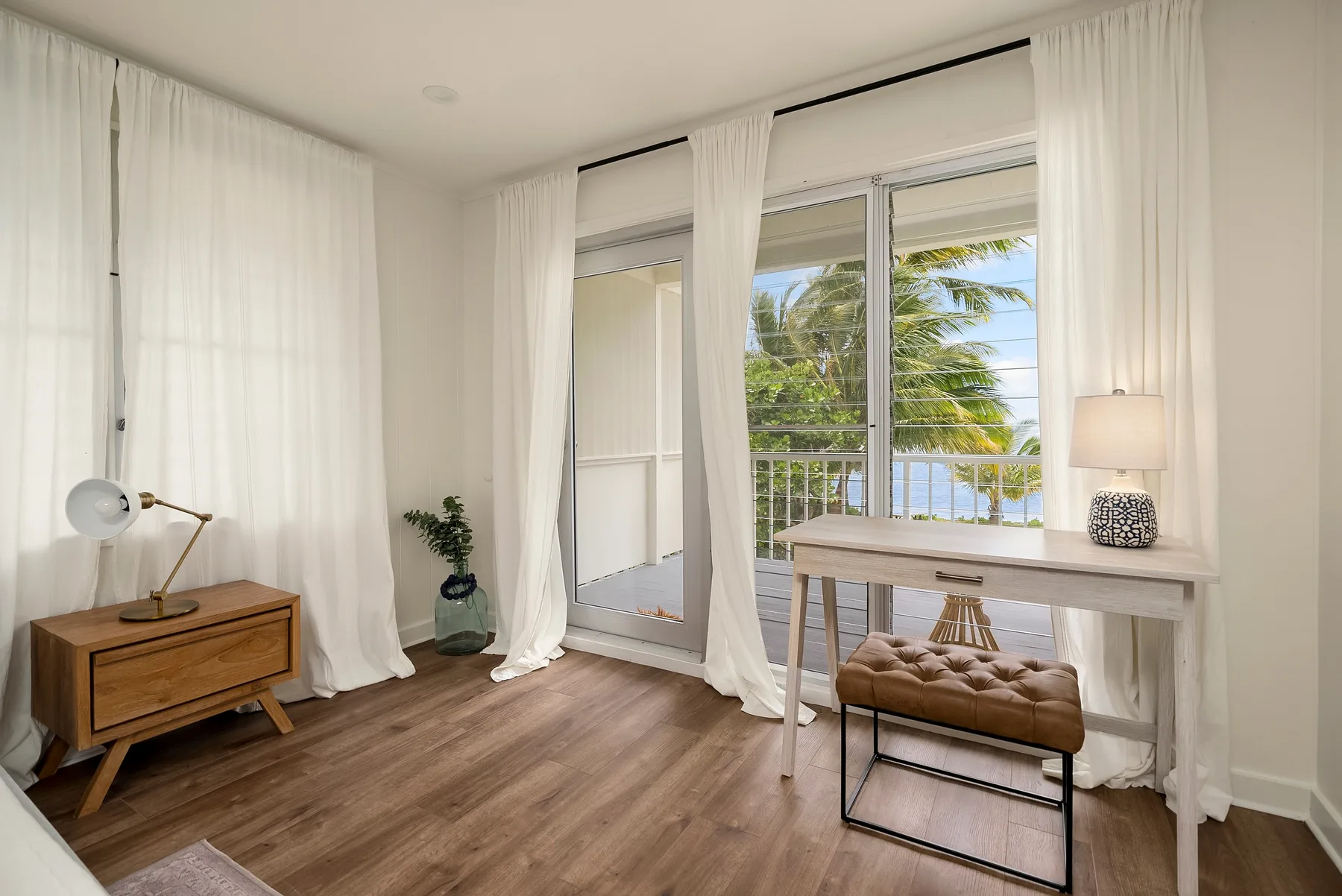 Una habitación luminosa con escritorio, cortinas largas y ventanas con vista a palmeras.