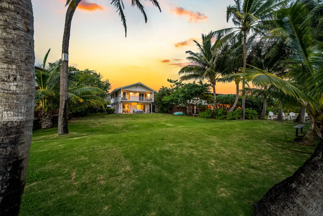 Una casa se alza tras un amplio césped, rodeada de palmeras al atardecer.