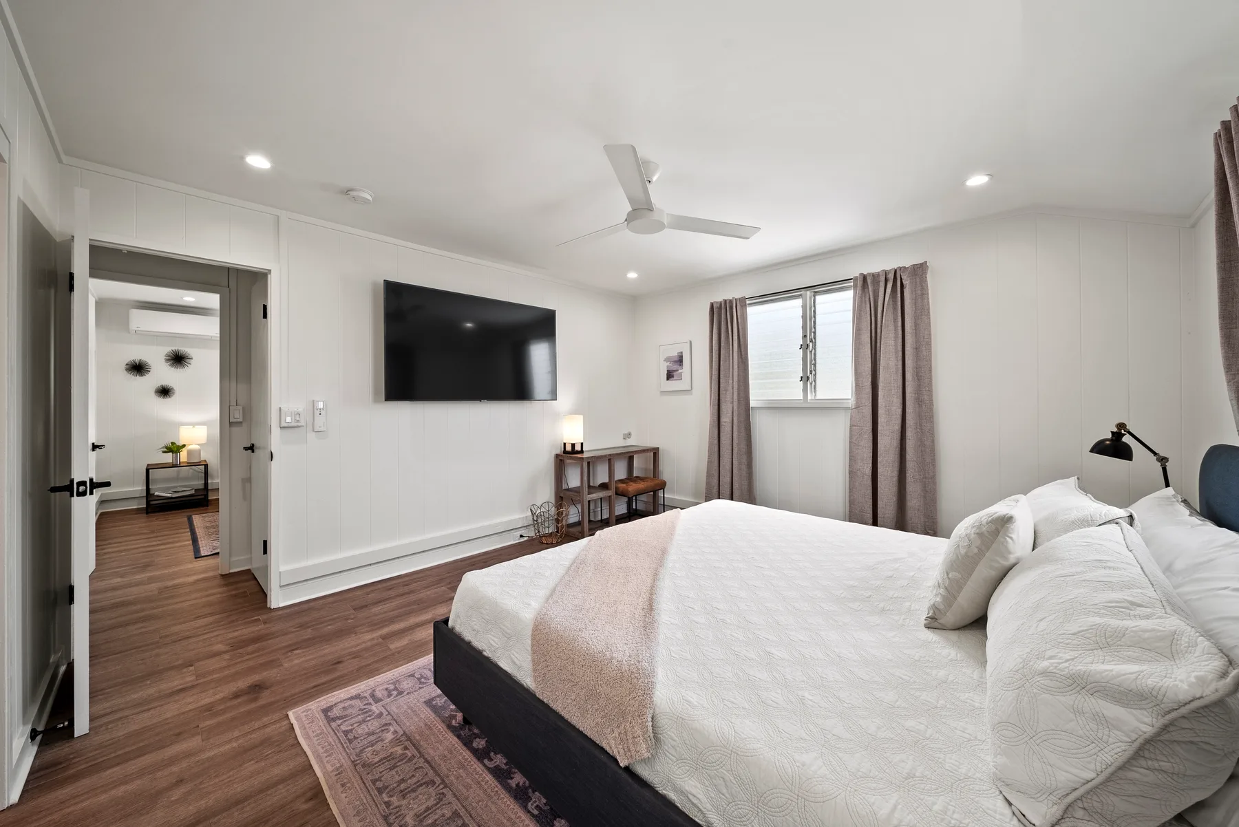 Un dormitorio luminoso con una cama, un televisor en la pared y una ventana con cortinas.