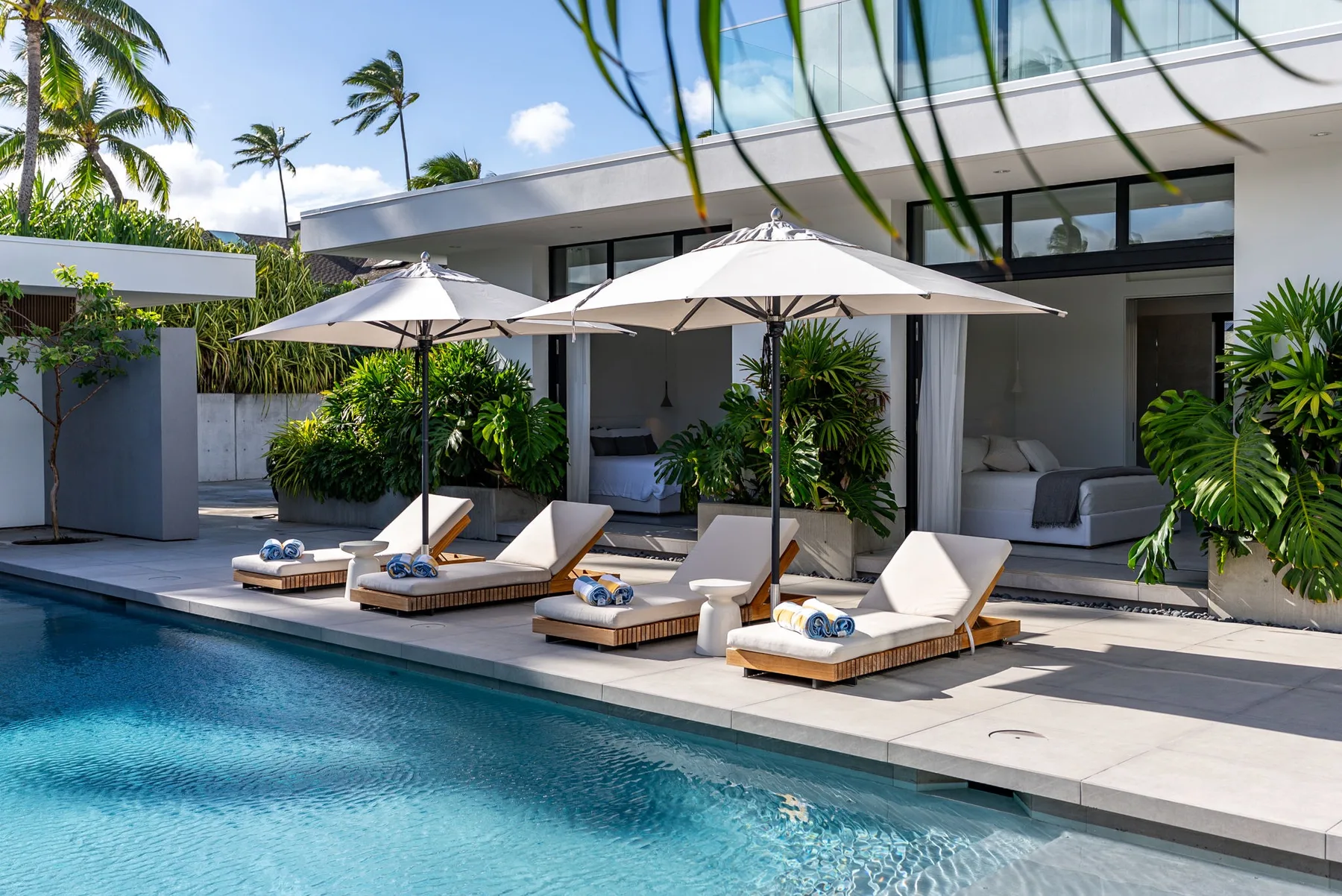 A swimming pool borders a patio with loungers and umbrellas beside a modern house.
