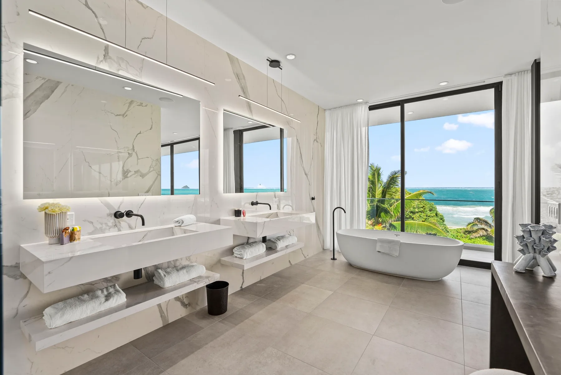 A bright bathroom has a freestanding tub beside floor-to-ceiling windows overlooking the ocean.