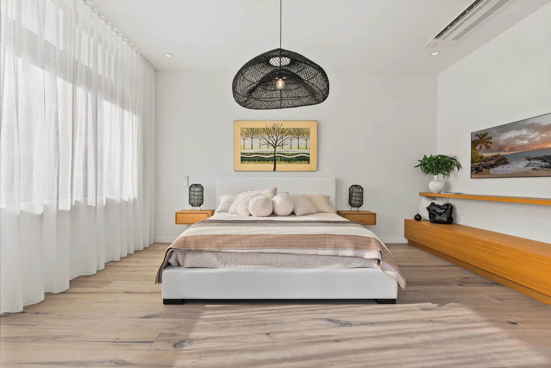 A bright bedroom with a bed beneath framed artwork and a woven pendant light.