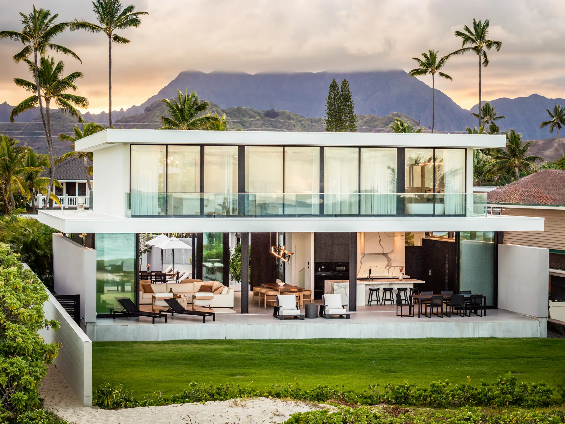A modern two-story house with glass windows overlooks a green lawn and mountain scenery.