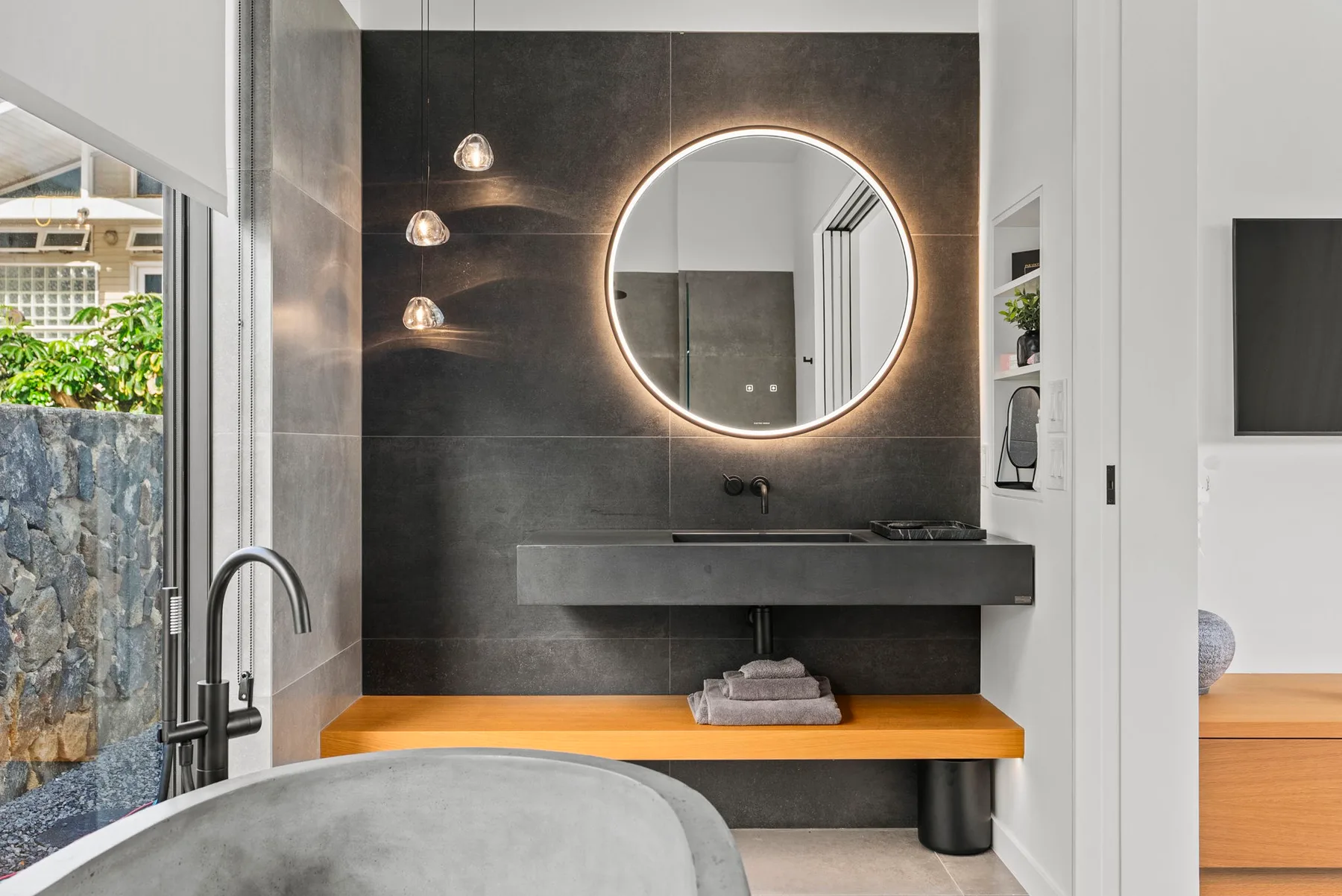A modern bathroom vanity with a round illuminated mirror and dark tiled wall.