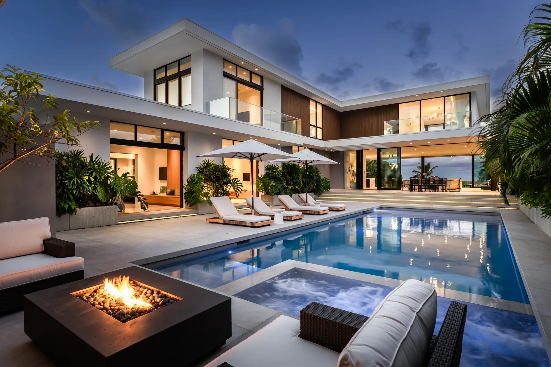 A modern house surrounds a swimming pool and patio seating at dusk.