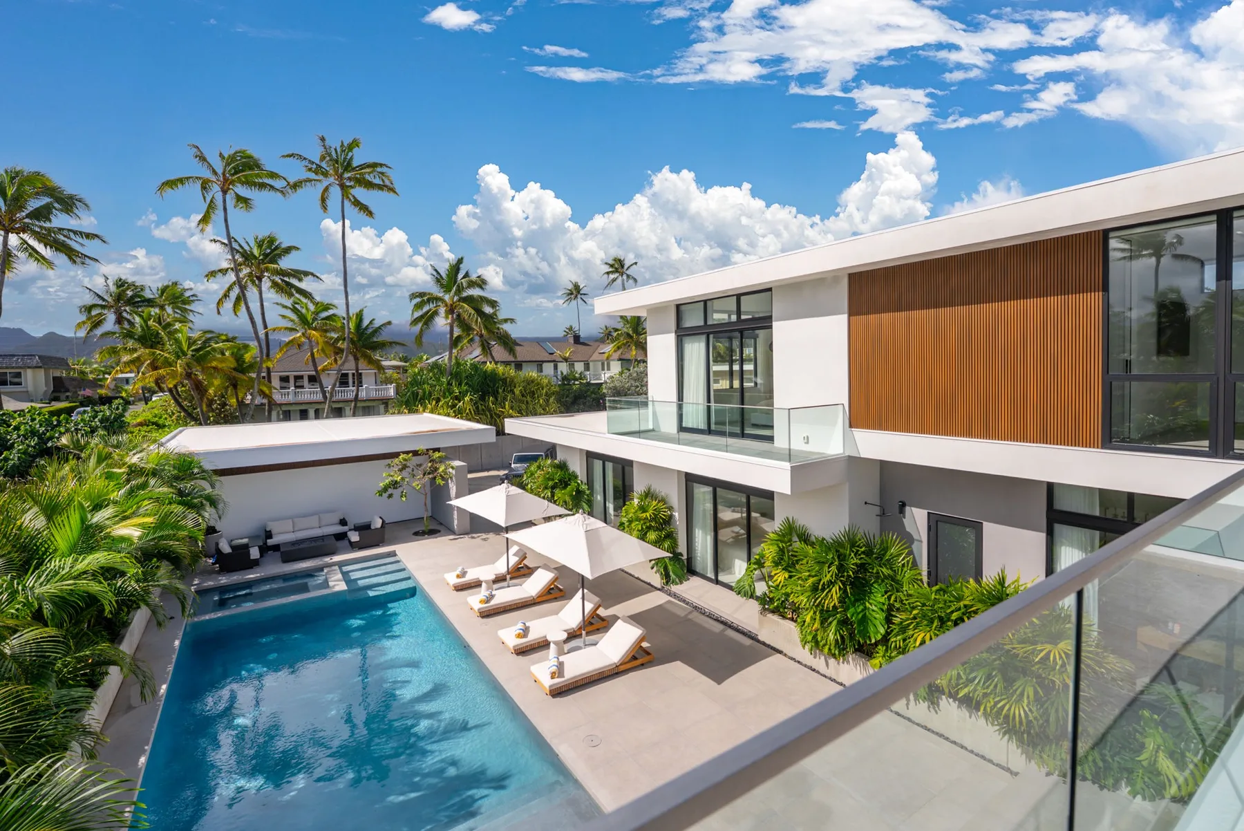 A modern white house overlooks a swimming pool and sun loungers amid tropical landscaping.
