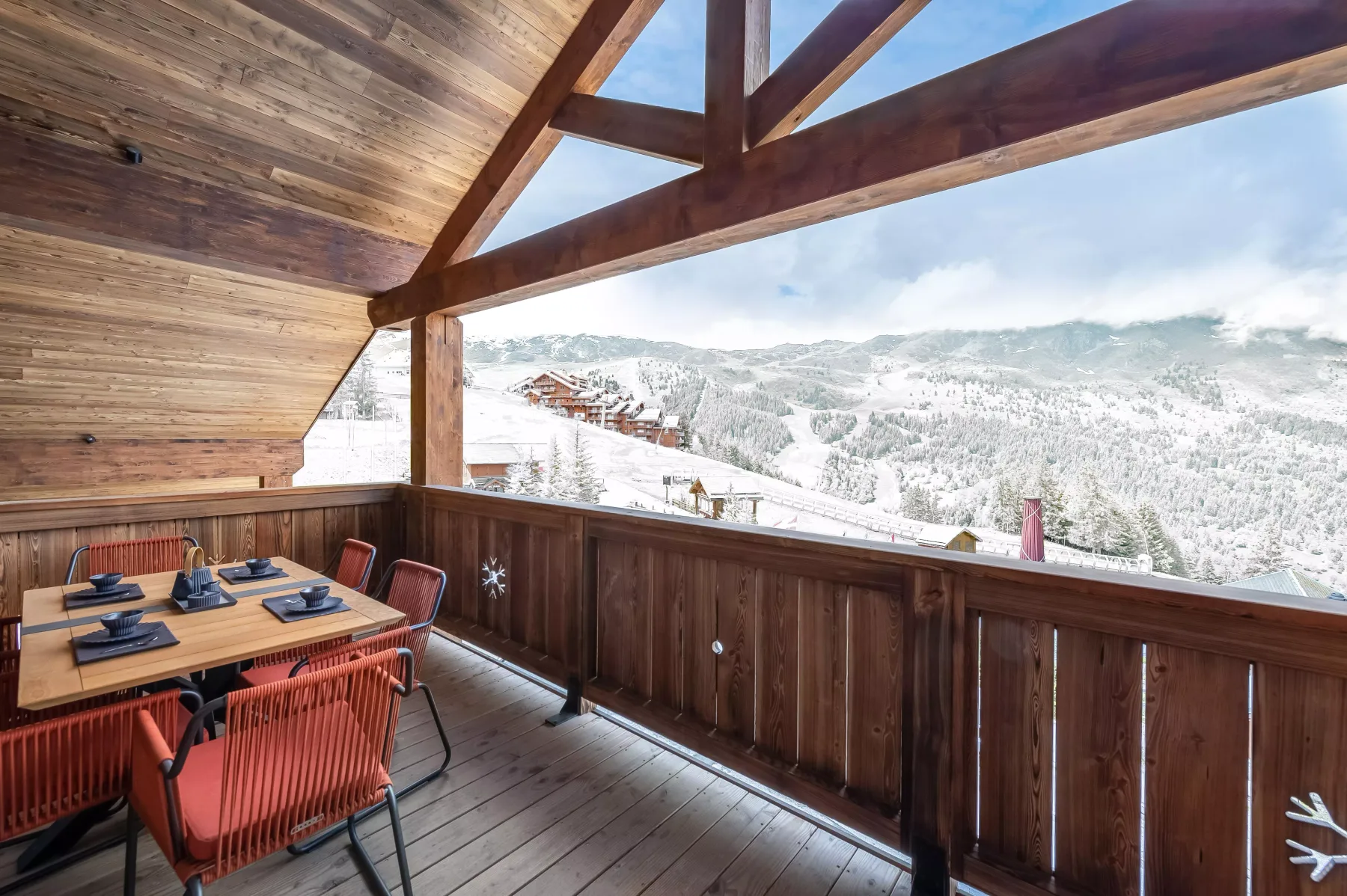 A covered wooden balcony has outdoor seating and views of snowy mountains.