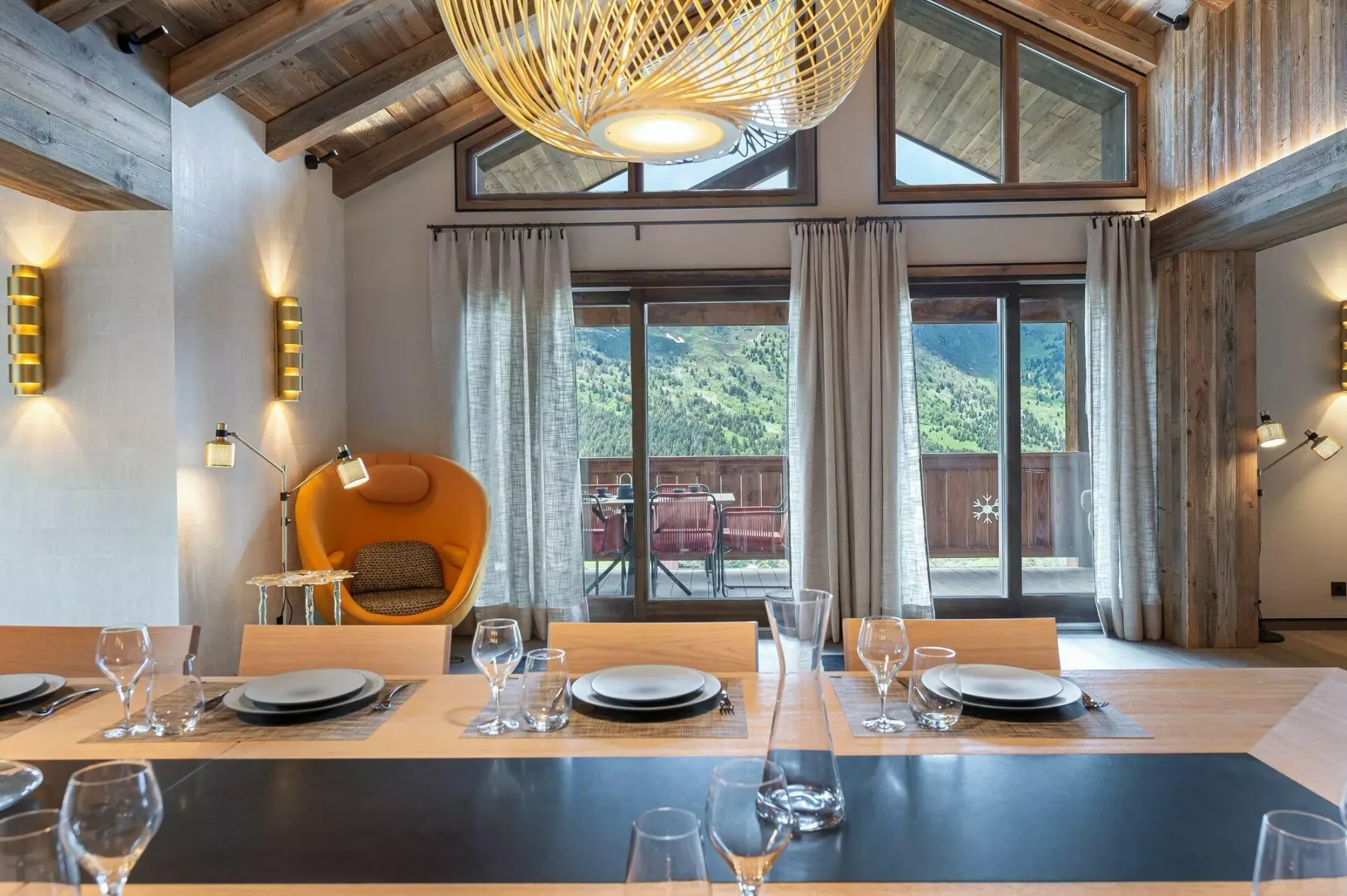 A dining area with a long table, pendant light, and windows overlooking a wooded view.