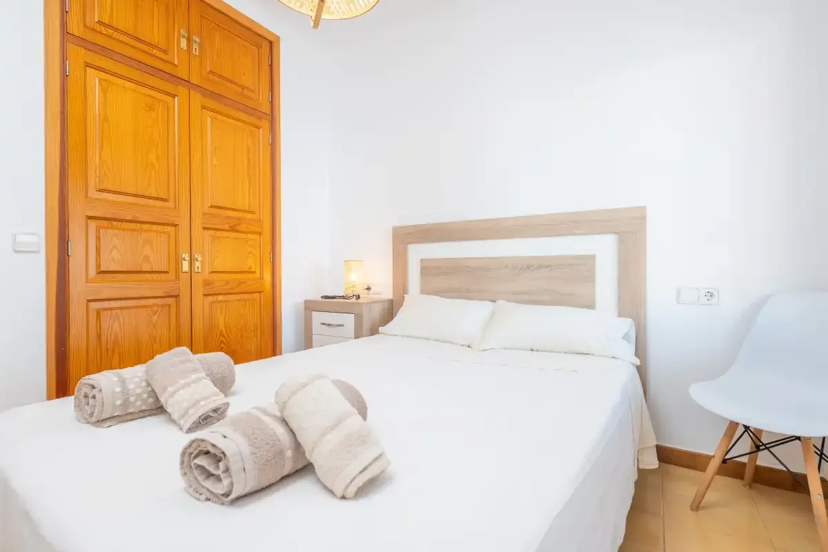 Un dormitorio luminoso con una cama, un armario de madera y una silla blanca.
