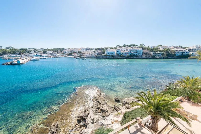 Bahía costera de aguas turquesas y claras, costa rocosa y edificios al otro lado.
